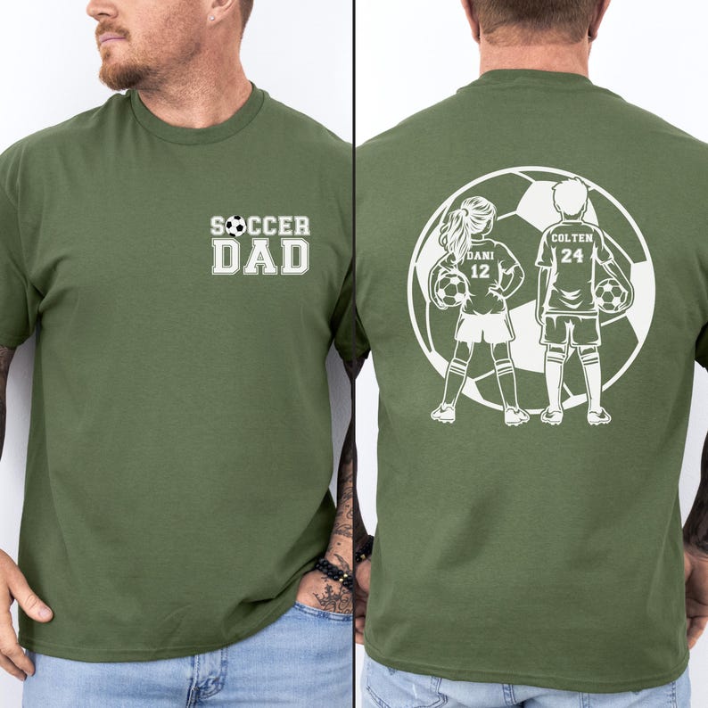 May include: Olive green t-shirt with the text "SOCCER DAD" in white on the front. The back features a white graphic of two soccer players inside a soccer ball, with the names "DANI 12" and "COLTEN 24".