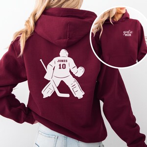 Op de afbeelding: Bordeaux hoodie met een witte hockey goalie graphic op de rug, met de naam "JONES" en het nummer "10". De voorkant van de hoodie heeft de tekst "goalie MOM" in het wit. Een comfortabele, casual top.