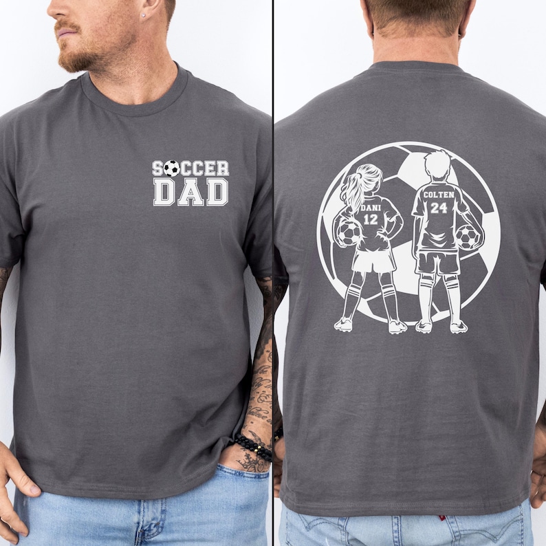 May include: A charcoal gray t-shirt with the text "SOCCER DAD" in white on the front. The back of the shirt features a white graphic of two soccer players inside a soccer ball, with the names "DANI 12" and "COLTEN 24".