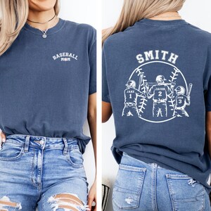 Puede incluir: Una camiseta azul marino con el texto "Baseball Mom" en la parte delantera y un dise&ntilde;o de tem&aacute;tica de b&eacute;isbol en la parte posterior. El dise&ntilde;o de la parte posterior presenta a tres jugadores de b&eacute;isbol con los nombres "Jake", "Jacob" y "John" y sus n&uacute;meros correspondientes 1, 2 y 3. El texto "Smith" est&aacute; por encima del dise&ntilde;o.