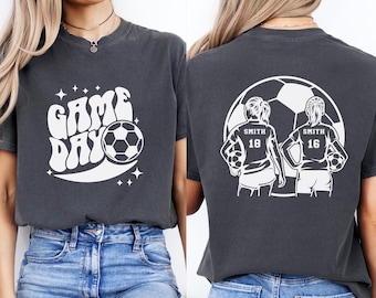 Könnte beinhalten: Zwei dunkelgraue T-Shirts mit weißen Fußball-Motiven. Das vordere Shirt trägt die Aufschrift "GAME DAY" mit einem Fußball. Das hintere Shirt zeigt zwei Fußballspielerinnen mit dem Text "SMITH 18" und "SMITH 16".