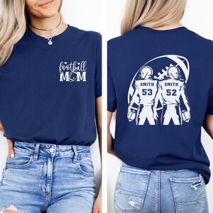 May include: Navy blue t-shirt with white graphic text "football MOM" on the front. The back features a football design with two football players, the name "SMITH" and the numbers "53" and "52".
