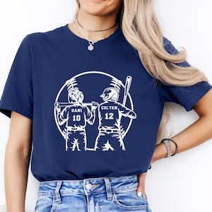 Op de afbeelding: Een donkerblauw t-shirt met een witte afbeelding van twee honkballers in een cirkel. De spelers staan ​​met hun ruggen naar elkaar en houden knuppels vast. De tekst "Dani 10" en "Colten 12" staat op de achterkant van hun shirts.