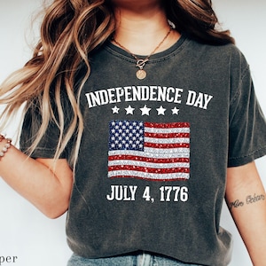 Może przedstawiać: Ciemnoszara koszulka z napisem "INDEPENDENCE DAY" nad błyszczącą grafiką amerykańskiej flagi. Poniżej flagi znajduje się napis "JULY 4, 1776". Koszulka ma swobodny styl.