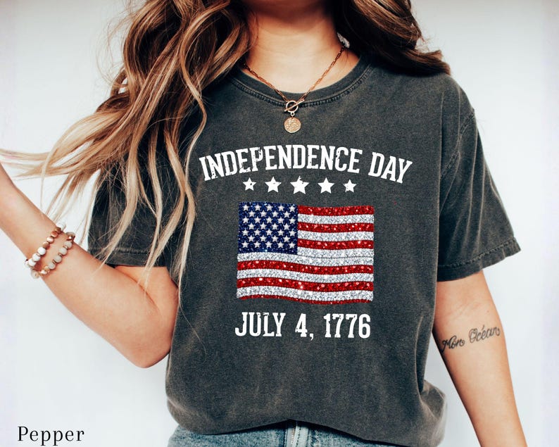 Może przedstawiać: Ciemnoszara koszulka z napisem "INDEPENDENCE DAY" nad błyszczącą grafiką amerykańskiej flagi. Poniżej flagi znajduje się napis "JULY 4, 1776". Koszulka ma swobodny styl.