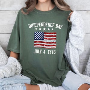 Może przedstawiać: Zielona koszulka na Dzień Niepodległości z postarzaną amerykańską flagą. Tekst "INDEPENDENCE DAY JULY 4, 1776" jest wydrukowany nad i pod flagą. Koszulka ma luźny kr&oacute;j.