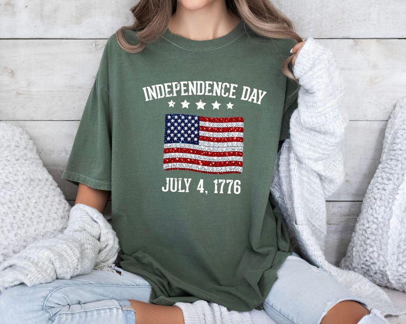 Może przedstawiać: Zielona koszulka na Dzień Niepodległości z postarzaną amerykańską flagą. Tekst "INDEPENDENCE DAY JULY 4, 1776" jest wydrukowany nad i pod flagą. Koszulka ma luźny kr&oacute;j.