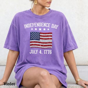 Może przedstawiać: Fioletowa koszulka z nadrukiem amerykańskiej flagi. Koszulka ma napis "INDEPENDENCE DAY" nad flagą i "JULY 4, 1776" pod nią. Koszulka jest z kr&oacute;tkim rękawem i ma swobodny styl.