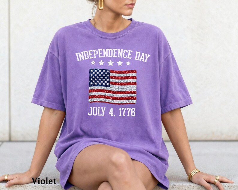 Może przedstawiać: Fioletowa koszulka z nadrukiem amerykańskiej flagi. Koszulka ma napis "INDEPENDENCE DAY" nad flagą i "JULY 4, 1776" pod nią. Koszulka jest z kr&oacute;tkim rękawem i ma swobodny styl.