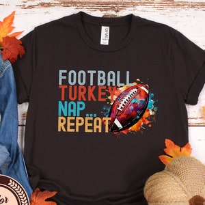 Könnte beinhalten: Ein schwarzes T-Shirt mit einem farbenfrohen Grafikdesign, das einen Fußball, die Worte "Football, Turkey, Nap, Repeat" und einen Aquarelleffekt zeigt.