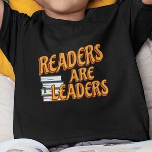 Puede incluir: Una persona joven con una camiseta negra con el texto "Readers Are Leaders" en naranja y blanco. La persona está sentada en un sofá sosteniendo un libro abierto.