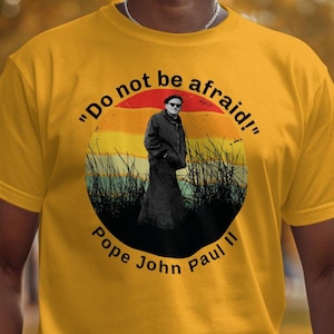 Puede incluir: Una camiseta amarilla con un gráfico de estilo vintage del Papa Juan Pablo II. El gráfico presenta una cita del Papa: "¡No tengan miedo!"