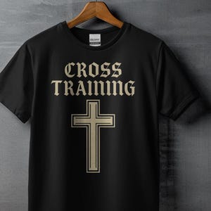 May include: Black t-shirt with the words "CROSS TRAINING" in a serif font above a cross graphic. The cross and text are in a light tan color. The shirt is hanging on a wooden hanger.