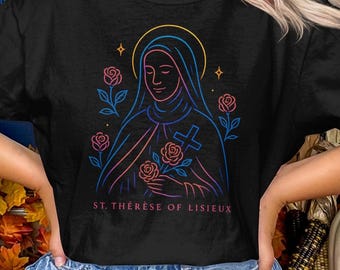 May include: Black t-shirt featuring a neon illustration of St. Thérèse of Lisieux. The design includes a halo, cross, roses, and the text "ST. THÉRÈSE OF LISIEUX". The artwork is rendered in shades of pink, blue, and yellow.