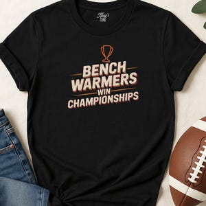 May include: Black t-shirt with the text "Bench Warmers Win Championships" in orange and white. The shirt features a trophy graphic above the text. A football is visible in the bottom right corner. The shirt is folded at the sleeves.