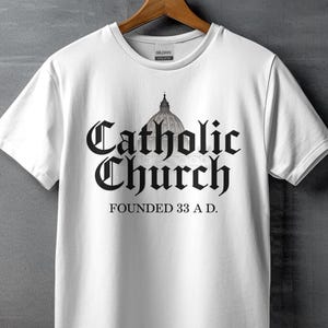 May include: White t-shirt featuring a black graphic design that reads "Catholic Church Founded 33 A.D." with an illustration of a church dome.
