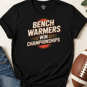May include: Black t-shirt with the text "BENCH WARMERS WIN CHAMPIONSHIPS" in white and orange lettering. The shirt has a crew neck and short sleeves. A football is visible in the bottom right corner.