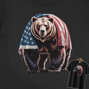 May include: A black t-shirt featuring a brown bear draped in an American flag. The bear is the central focus, with the flag's red and white stripes and blue star field prominently displayed. A smaller image of the design is on a t-shirt on a hanger.