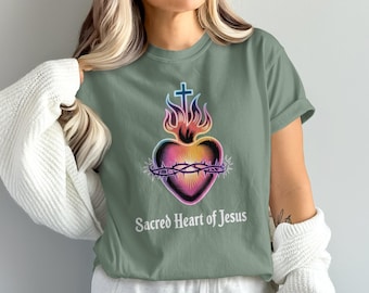 May include: Sage green t-shirt featuring a colorful Sacred Heart of Jesus graphic. The design includes a heart, a crown of thorns, flames, and a cross. The text "Sacred Heart of Jesus" is printed below the graphic in a white, sans-serif font.