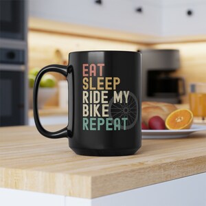 May include: Black coffee mug with a white graphic that reads "Eat Sleep Ride My Bike Repeat" with a bicycle wheel graphic. The mug is on a wooden surface.