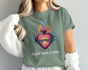 Könnte beinhalten: Salbeigrünes T-Shirt mit einer Grafik eines Herzens, einer Dornenkrone, Flammen und einem Kreuz. Das Herz ist rosa mit gelben und orangen Akzenten. Der Text "Sacred Heart of Jesus" steht darunter.