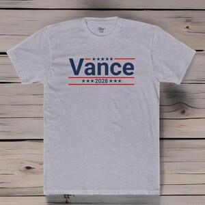 May include: A gray t-shirt with a blue and red design. The design features the word "Vance" in blue with red stars and stripes above and below the word. The text "2028" is also included in the design.