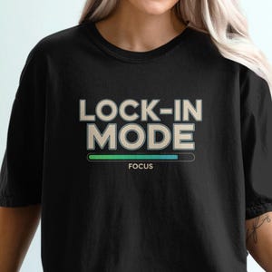 May include: Black t-shirt with the text "LOCK-IN MODE" in a light beige font, with a green and blue progress bar and the word "FOCUS" below. The shirt is a solid black color.