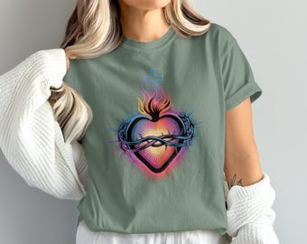 May include: Sage green t-shirt featuring a vibrant graphic of a heart with flames, a crown of thorns, and a cross. The heart design incorporates a spectrum of colors, including pink, orange, and blue, creating a striking visual effect. The shirt has short sleeves.