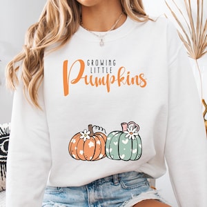 May include: White sweatshirt with the text "Growing Little Pumpkins" in orange script. The sweatshirt features two illustrated pumpkins, one orange with white flowers and the other green with white hearts and a white flower.