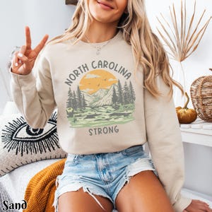 Op de afbeelding: Een beige sweatshirt met een grafisch ontwerp van een bergketen met zonsondergang en de tekst "North Carolina Strong".