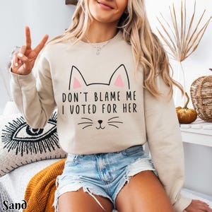 May include: A beige sweatshirt with a cat graphic and the text "Don't blame me I voted for her".