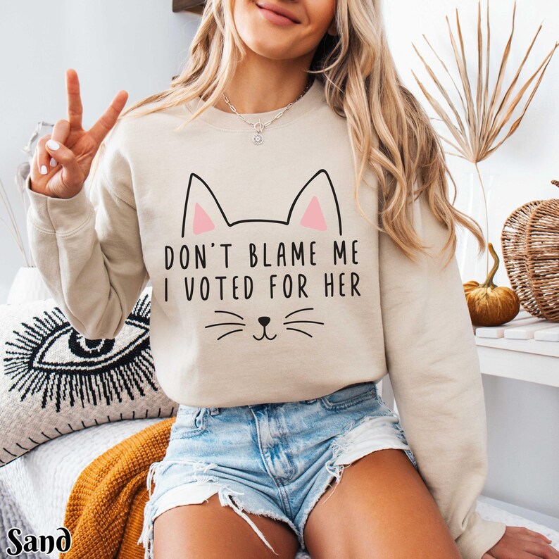 May include: A beige sweatshirt with a cat graphic and the text "Don't blame me I voted for her".