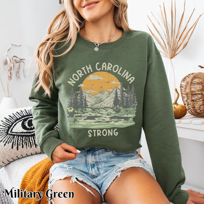 Op de afbeelding: Een groen sweatshirt met een vintage-stijl grafische afbeelding van een bergketen en een zonsondergang. De afbeelding zegt "North Carolina Strong".