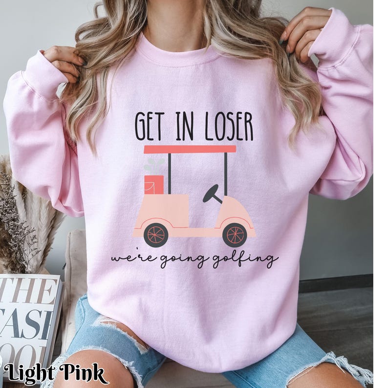 K&ouml;nnte beinhalten: Ein rosa Sweatshirt mit dem Text "Get in loser we're going golfing" und einem Cartoon-Golfwagen.