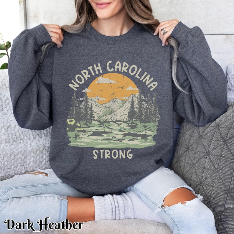 Op de afbeelding: Een donker grijs sweatshirt met een vintage-stijl afbeelding van een bergketen en een ondergaande zon. De tekst "North Carolina Strong" is onder de afbeelding gedrukt.