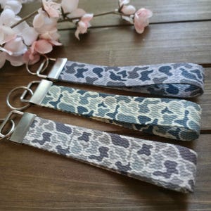 May include: Three camouflage-patterned key fobs in shades of gray, blue, and white. Each fob has a silver-colored metal clasp and key ring. The key fobs are arranged on a wooden surface, with pink flowers in the background.