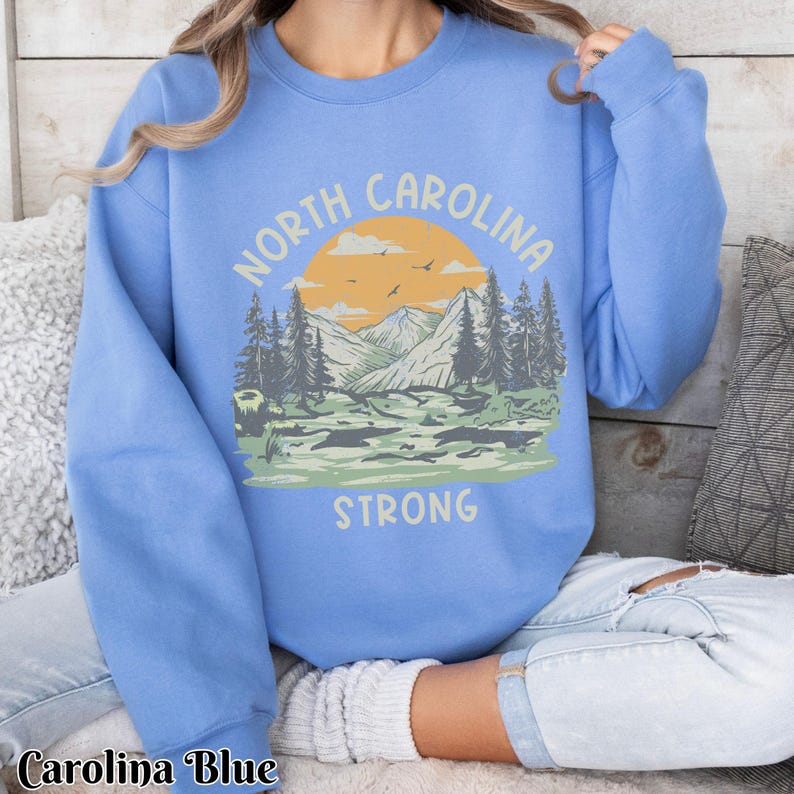 Op de afbeelding: Een lichtblauw sweatshirt met een grafisch ontwerp van een bergketen met zonsondergang en de tekst "NORTH CAROLINA STRONG".