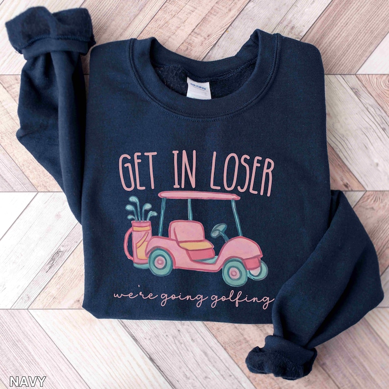Puede incluir: Sudadera azul marino con el texto "GET IN LOSER" sobre una ilustraci&oacute;n de un carrito de golf rosa. Debajo, el texto dice "we're going golfing". La sudadera tiene cuello redondo y mangas largas.