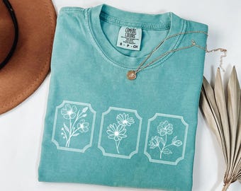 May include: A teal blue t-shirt with a white floral design. The shirt has a crew neck and short sleeves. The text "COMFORT COLORS" is printed on the tag.