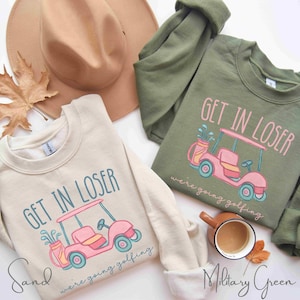 Puede incluir: Dos sudaderas, una color arena y otra verde militar, cada una con un dise&ntilde;o de carrito de golf y el texto "GET IN LOSER we're going golfing." Un sombrero beige y una taza de caf&eacute; tambi&eacute;n est&aacute;n en la imagen.