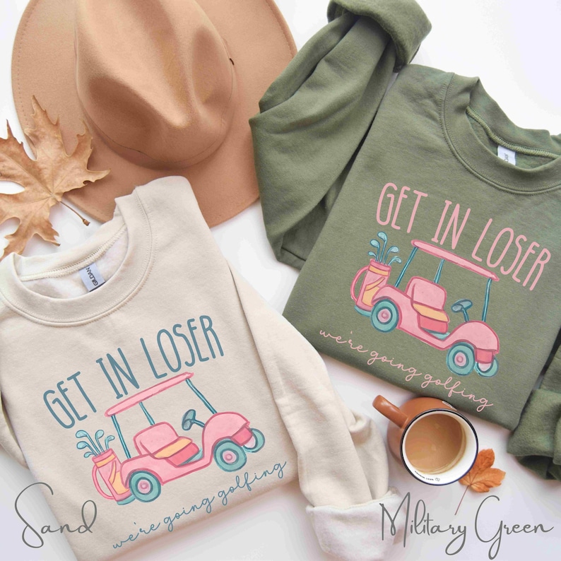 Puede incluir: Dos sudaderas, una color arena y otra verde militar, cada una con un dise&ntilde;o de carrito de golf y el texto "GET IN LOSER we're going golfing." Un sombrero beige y una taza de caf&eacute; tambi&eacute;n est&aacute;n en la imagen.