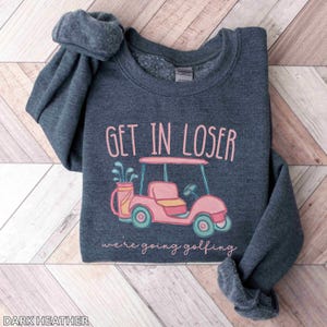 Puede incluir: Sudadera gris oscuro con el texto "GET IN LOSER" sobre una ilustraci&oacute;n de carrito de golf rosa. Debajo del carrito, el texto dice "we're going golfing". La sudadera est&aacute; doblada sobre una superficie de madera clara.