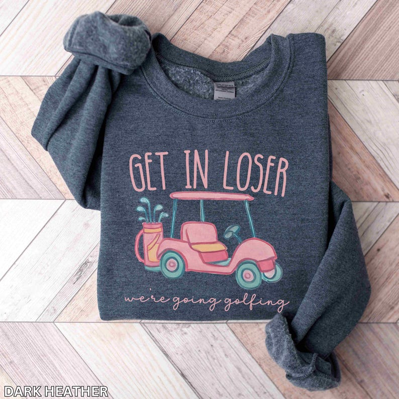 Puede incluir: Sudadera gris oscuro con el texto "GET IN LOSER" sobre una ilustraci&oacute;n de carrito de golf rosa. Debajo del carrito, el texto dice "we're going golfing". La sudadera est&aacute; doblada sobre una superficie de madera clara.
