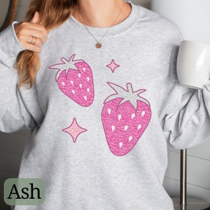 May include: A gray sweatshirt with a pink glitter strawberry design and two pink glitter stars. The sweatshirt is labeled "Ash" in the bottom left corner.