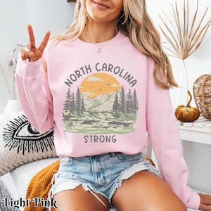 Op de afbeelding: Een lichtroze sweatshirt met een grafisch ontwerp van een bergketen met zonsondergang en de tekst "North Carolina Strong".