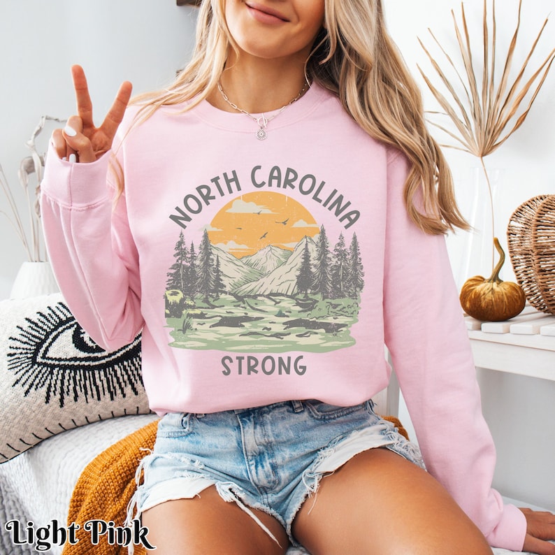 Op de afbeelding: Een lichtroze sweatshirt met een grafisch ontwerp van een bergketen met zonsondergang en de tekst "North Carolina Strong".