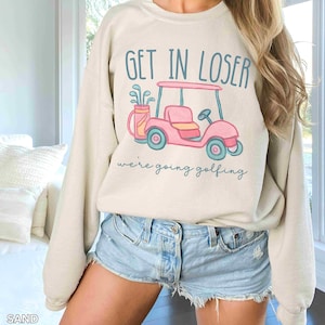 Puede incluir: Sudadera color arena con el texto "GET IN LOSER" sobre un gr&aacute;fico de carrito de golf rosa. Debajo del carrito, el texto dice "we're going golfing". El carrito de golf tiene ruedas y detalles turquesa.