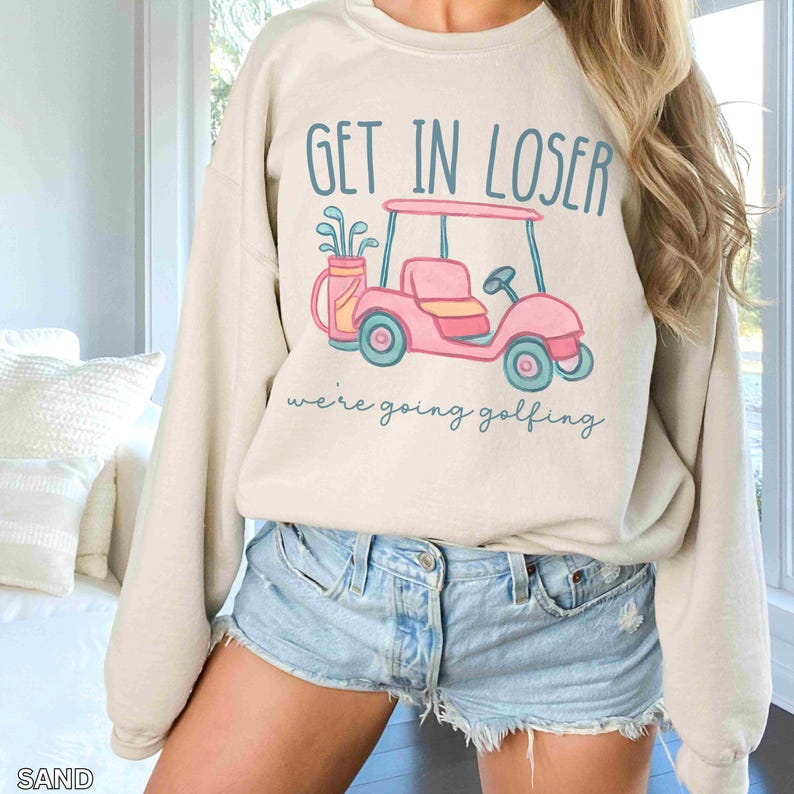 Puede incluir: Sudadera color arena con el texto "GET IN LOSER" sobre un gr&aacute;fico de carrito de golf rosa. Debajo del carrito, el texto dice "we're going golfing". El carrito de golf tiene ruedas y detalles turquesa.