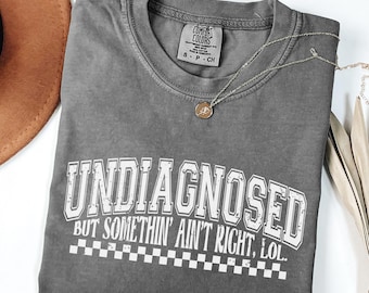 May include: Grey t-shirt with a white graphic that reads "UNDIAGNOSED BUT SOMETHIN' AIN'T RIGHT, LOL..." with a chequered pattern below the text.
