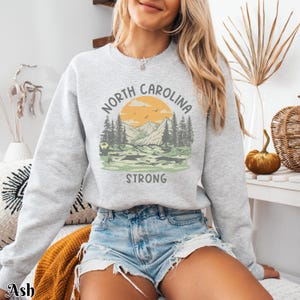 Op de afbeelding: Een grijs sweatshirt met een grafische print van een berglandschap en de tekst "North Carolina Strong".